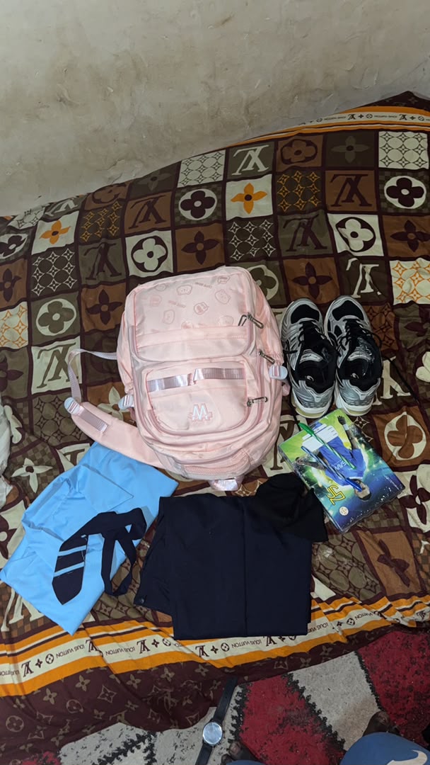 A blue shirt, pink school bag, black shoes, and school supplies laid out on a patterned mat.